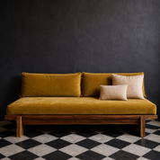 A modern wooden daybed with a mustard yellow velvet cushioned seat and back, styled with two beige throw pillows against a dark textured wall and a black-and-white checkered floor.