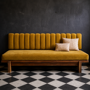 A mustard yellow velvet daybed with a channel-tufted backrest and wooden legs, styled with two beige textured throw pillows against a dark textured wall and a black-and-white checkered floor.