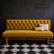 A mustard yellow tufted velvet daybed with a wooden frame and two beige accent pillows, set against a dark textured wall on a black and white checkered floor.