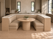 A U-shaped upholstered banquette seating unit with a round wooden pedestal table, set in a modern kitchen with light wood cabinetry and warm natural light.