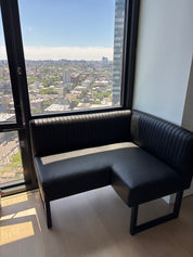 A black corner booth bench with ribbed upholstery and a high back, positioned next to a large window with a city view.